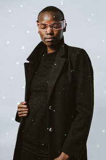 Porträt einer selbstbewussten jungen, mutigen afroamerikanischen Frau mit einer intelligenten, futuristischen VR-Brille und einem langen schwarzen Trenchcoat, die mit geschlossenen Augen vor einem mit Neonlicht beleuchteten Hintergrund steht - ADSF52138