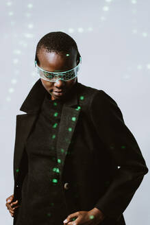 Porträt einer selbstbewussten jungen, mutigen afroamerikanischen Frau mit einer intelligenten, futuristischen VR-Brille und einem langen schwarzen Trenchcoat, die mit geschlossenen Augen vor einem mit Neonlicht beleuchteten Hintergrund steht - ADSF52139