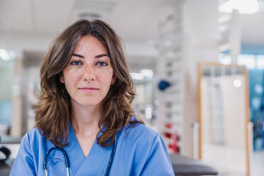 Porträt einer jungen Ärztin mit Stethoskop um den Hals und in Uniform, die in die Kamera schaut, während sie an einem Tisch in einem Krankenhaus sitzt, vor einem unscharfen Hintergrund in beleuchtetem Licht - ADSF52149