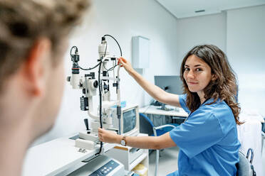 Lächelnde junge Ärztin in Uniform und Stethoskop, die in die Kamera blickt, während sie mit der Hand auf dem Spaltlampen-Biomikroskop in einem Krankenhaus unter beleuchtetem Licht steht und auf einen unscharfen anonymen Mann zeigt - ADSF52152