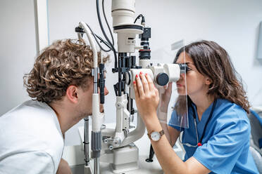 Seitenansicht einer jungen Ärztin in medizinischer Uniform und mit Stethoskop, die wegschaut, während sie mit einem anonymen männlichen Patienten an einem Spaltlampen-Biomikroskop sitzt, während einer Untersuchung in einer Klinik in beleuchtetem Licht - ADSF52153