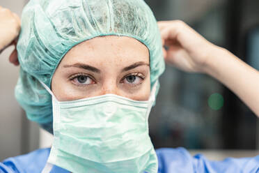 Junge Chirurgin in Uniform mit medizinischer Maske und Kappe schaut in die Kamera vor dem unscharfen Hintergrund eines modernen Krankenhauses - ADSF52158
