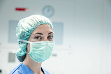 Seitenansicht einer Chirurgin mit grüner Kappe und Schutzmaske, die in einem modernen Krankenhaus in die Kamera schaut - ADSF52160