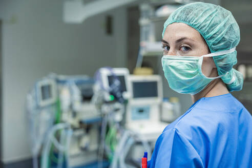 Professional female surgeon in uniform and protective mask with cap standing against blurred interior of operating room while looking at camera - ADSF52161