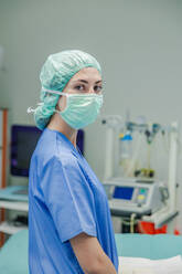 Professional female surgeon in uniform and protective mask with cap standing against blurred interior of operating room while looking at camera - ADSF52162