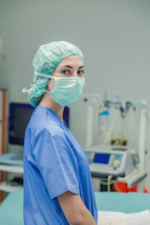 Professionelle weibliche Chirurgin in Uniform und Schutzmaske mit Kappe steht vor dem unscharfen Inneren eines Operationssaals und schaut in die Kamera - ADSF52162