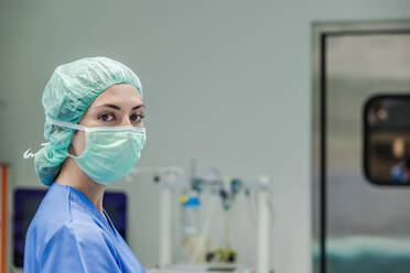 Professionelle weibliche Chirurgin in Uniform und Schutzmaske mit Kappe steht vor dem unscharfen Inneren eines Operationssaals und schaut in die Kamera - ADSF52163