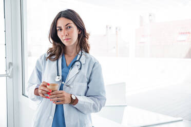 Young female doctor wearing medical uniform and stethoscope standing in modern hospital with cup while looking at camera against white background - ADSF52164
