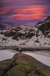 Ein einsamer Abenteurer steht mit einer Lampe in der Hand auf einem moosbewachsenen Felsvorsprung in Island, wo sich der Himmel in der Dämmerung in Rosa- und Violetttönen über der zerklüfteten, schneebedeckten Landschaft spiegelt. - ADSF52167