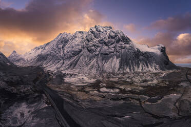 Ein malerischer Blick auf die majestätischen schneebedeckten Berge Islands bei Sonnenuntergang mit einer kurvenreichen Straße, die durch die zerklüftete Landschaft führt. - ADSF52174