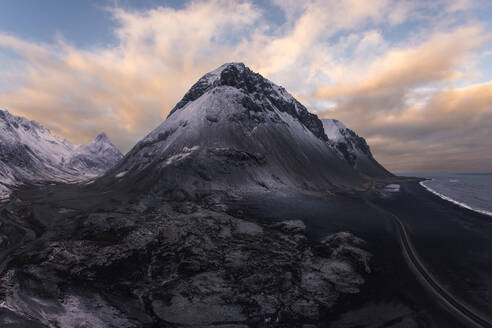 Eine atemberaubende Ansicht, die die raue Schönheit einer isländischen Landschaft mit einem schneebedeckten Berg neben einer dramatischen Küstenlinie unter einem Dämmerungshimmel einfängt. - ADSF52176