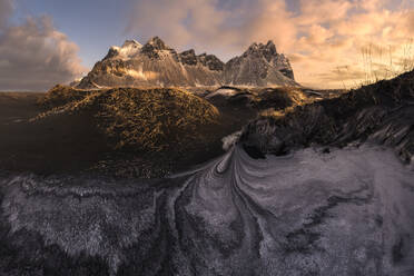 Eine atemberaubende isländische Landschaft mit majestätischen Berggipfeln, die in das warme Licht des Sonnenaufgangs getaucht sind, kontrastiert mit dunklem Vulkansand und zarten Frostmustern. - ADSF52177