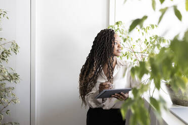 Nachdenkliche junge afroamerikanische Geschäftsfrau mit Faux-Locs-Frisur, die ein digitales Tablet und einen Stift hält, während sie durch ein Fenster im Büro schaut - ADSF52192