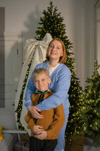 Eine herzliche Umarmung zwischen einer lächelnden Mutter und ihrem Sohn vor einem geschmückten Weihnachtsbaum, die die Freude der Weihnachtszeit zum Ausdruck bringt. - ADSF52194