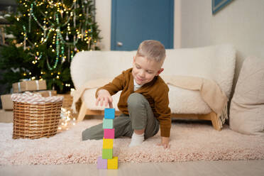 Ein kleiner Junge ist ganz in sein Spiel vertieft und stapelt bunte Blöcke auf einem weichen Teppich, während im Hintergrund ein Weihnachtsbaum leuchtet - ADSF52197