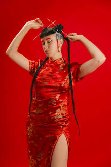 Eine Frau steht selbstbewusst in einem leuchtend roten, traditionellen chinesischen Kleid mit goldenen Verzierungen vor einem passenden roten Hintergrund. - ADSF52207