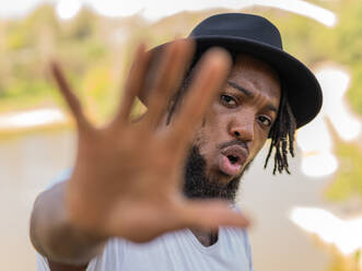 Young African American bearded man with dreadlocks in black hat outstretching hand towards camera and showing stop gesture - ADSF52223