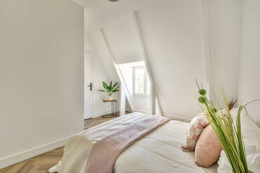 Comfortable bed with pillows and cushions with blanket in white loft bedroom with window and plant - ADSF52239