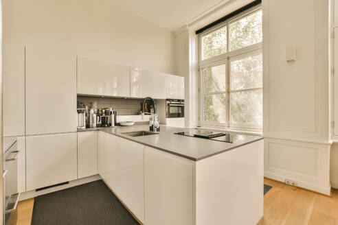 Sink and induction at countertop of modern kitchen of luxurious house with large window and white cabinets - ADSF52255