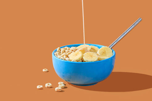 A blue bowl of cereal with banana slices being served with a splash of milk on an orange background. - ADSF52263
