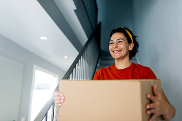 Fröhliche erwachsene Frau in Freizeitkleidung mit dunklen Haaren, die einen Karton auf der Treppe trägt, während sie tagsüber in eine neue Wohnung umzieht - ADSF52302