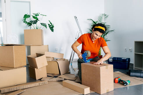 Positive female adult in red shirt and yellow hairband kneeling and packing carton box with tape dispenser during relocation - ADSF52309
