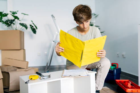Concentrated teenage boy in casual clothing kneeling and checking notes on clipboard before assembling furniture during relocation - ADSF52327