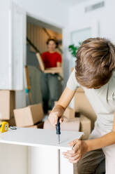 Crop unrecognizable boy using screwdriver while assembling wooden shelf during relocation against blurred mother in background - ADSF52328