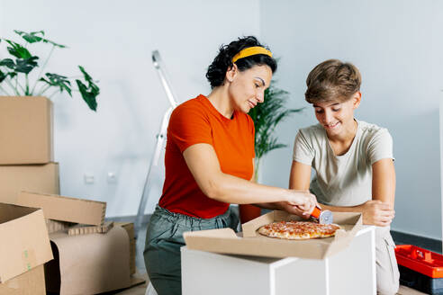 Glückliche Mutter und Sohn in legerer Kleidung schneiden zu Hause beim Auspacken der Sachen nach dem Umzug in eine neue Wohnung eine leckere Pizza auf - ADSF52329