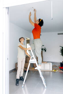 Seitenansicht einer Frau in Freizeitkleidung auf einer Leiter, die eine Lampe repariert, und eines Sohnes, der die Leiter hält und während des Umzugs nach oben schaut - ADSF52333