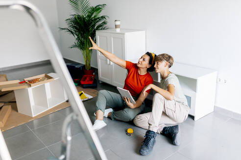 Hochwinklige Seitenansicht der Mutter, die mit ihrem Sohn zusammensitzt, der einen Notizblock hält und nach oben zeigt, während er in eine neue Wohnung zieht - ADSF52336