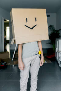 Gesichtslose Person in Freizeitkleidung, die Kopf und Schultern mit einem leeren Karton mit Gesichtsemoticon vor einem düsteren Hintergrund bedeckt - ADSF52344