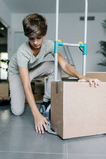 Focused brown haired teen in casual outfit kneeling and packing carton box on cart during moving into new home - ADSF52346