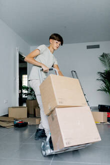 Side view of teen in gray casual clothes moving two carton boxes with cart during relocation to new house - ADSF52347