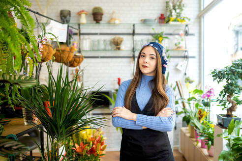 Junge Verkäuferin in Schürze steht mit gekreuzten Händen in einem Blumenladen in der Nähe verschiedener Topfpflanzen und Blumen während eines Arbeitstages - ADSF52352