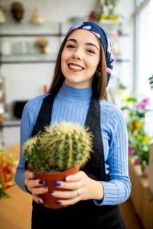 Positive junge Gärtnerin mit Kopftuch und Schürze, die in einem Blumenladen steht und bei der Arbeit einen getopften Kaktus hält - ADSF52354