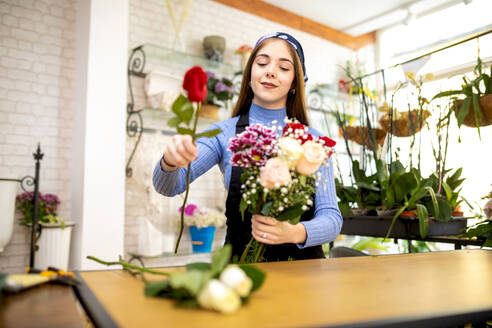 Positive junge Floristin beim Arrangieren eines Blumenstraußes aus Rosen, Chrysanthemen und Schleierkraut bei der Arbeit in einem Blumenladen - ADSF52358