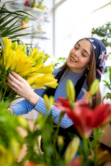 Lächelnde junge Floristin in Freizeitkleidung und Kopftuch arrangiert tagsüber frische gelbe Blumen bei der Arbeit in der Floristik - ADSF52361