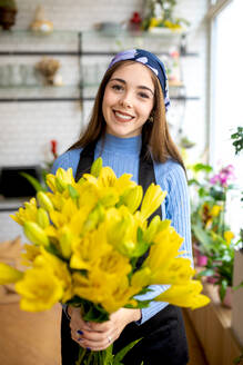 Lächelnde junge Floristin in Freizeitkleidung und Kopftuch, die frische gelbe Blumen hält, während sie tagsüber in der Floristik arbeitet - ADSF52362