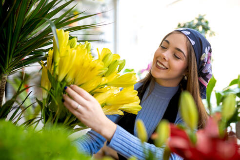 Lächelnde junge Floristin in Freizeitkleidung und Kopftuch arrangiert tagsüber frische gelbe Blumen bei der Arbeit in der Floristik - ADSF52363