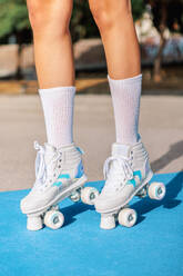 Crop black female legs in white socks and quad roller sneakers standing on blue and beige colored sports ground with shadow on sunny day - ADSF52406