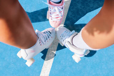 Top view of crop anonymous young black female athletes legs in white roller sneaker skates meeting at point while standing on blue sports ground on sunny day - ADSF52410