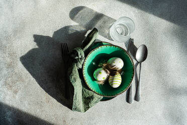 Top view of Easter table setting, showcasing a vibrant green ceramic plate with two decorative Easter eggs adorned with white and green patterns and delicate feathers, placed on gray surface between napkin and cutlery and glass of water - ADSF52413