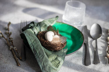 Hoher Winkel des Ostertischs, der einen leuchtend grünen Keramikteller mit einem kleinen Nest mit gesprenkelten Eiern und Federn zeigt, der auf einer grauen Fläche mit Serviette, Besteck und einem Glas Wasser steht - ADSF52414