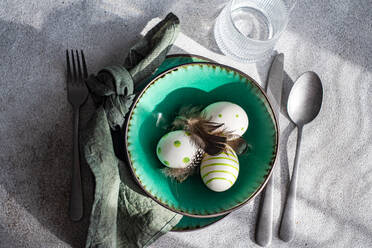 Top view of Easter table setting, showcasing a vibrant green ceramic plate with two decorative Easter eggs adorned with white and green patterns and delicate feathers, placed on gray surface between napkin and cutlery and glass of water - ADSF52416