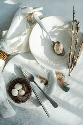 Draufsicht auf ein elegantes Osterarrangement mit makellosen weißen Tellern mit zartem Prägemuster und einem geflochtenen Nest mit gesprenkelten Eiern auf einem Teller, begleitet von Gabel und Messer, Federn und Zweigen - ADSF52417