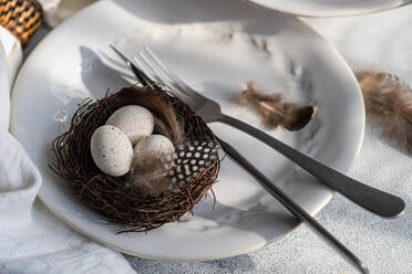 Hochformatige Darstellung eines eleganten Osterarrangements, bestehend aus einem makellosen weißen Teller mit einem zarten Prägemuster und einem geflochtenen Nest mit gesprenkelten Eiern, begleitet von Gabel und Messer und Federn - ADSF52418