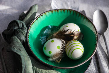 High angle closeup of Easter table setting, showcasing a vibrant green ceramic plate with two decorative Easter eggs adorned with white and green patterns and delicate feathers, placed on gray surface between napkin and cutlery and glass of water - ADSF52419