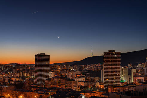 Die Mondsichel hängt am Dämmerungshimmel, während die Morgendämmerung über der Skyline der Innenstadt von Tiflis hereinbricht und die Stadt in ein warmes Licht taucht - ADSF52420