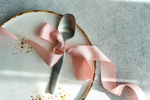 A top view of a shiny spoon tied with a soft pink ribbon on a ceramic plate with a golden rim, set against a textured background - ADSF52421